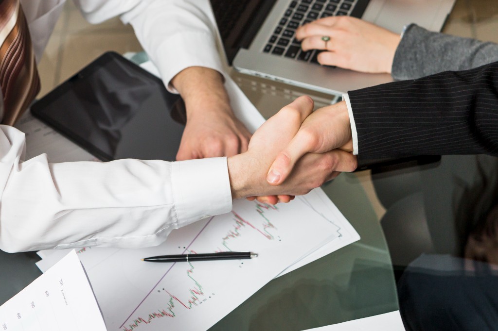 Top-down view of two professionals shaking hands over a glass desk with financial charts, a tablet, and a laptop