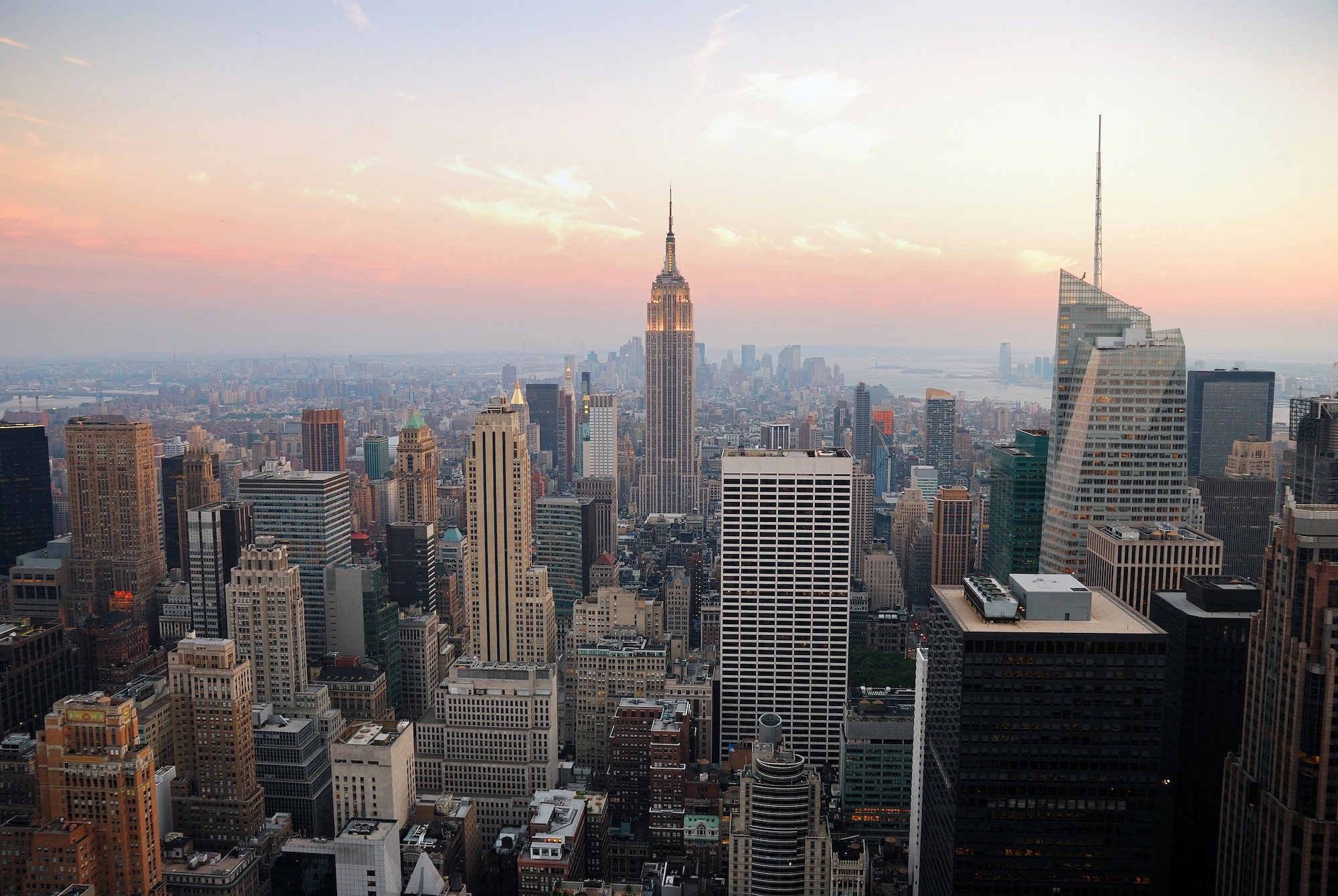 High-angle wide view south across Midtown Manhattan at golden hour, Empire State Building centered under a soft pink-orange sky, dense towers and street grids fading toward the harbor